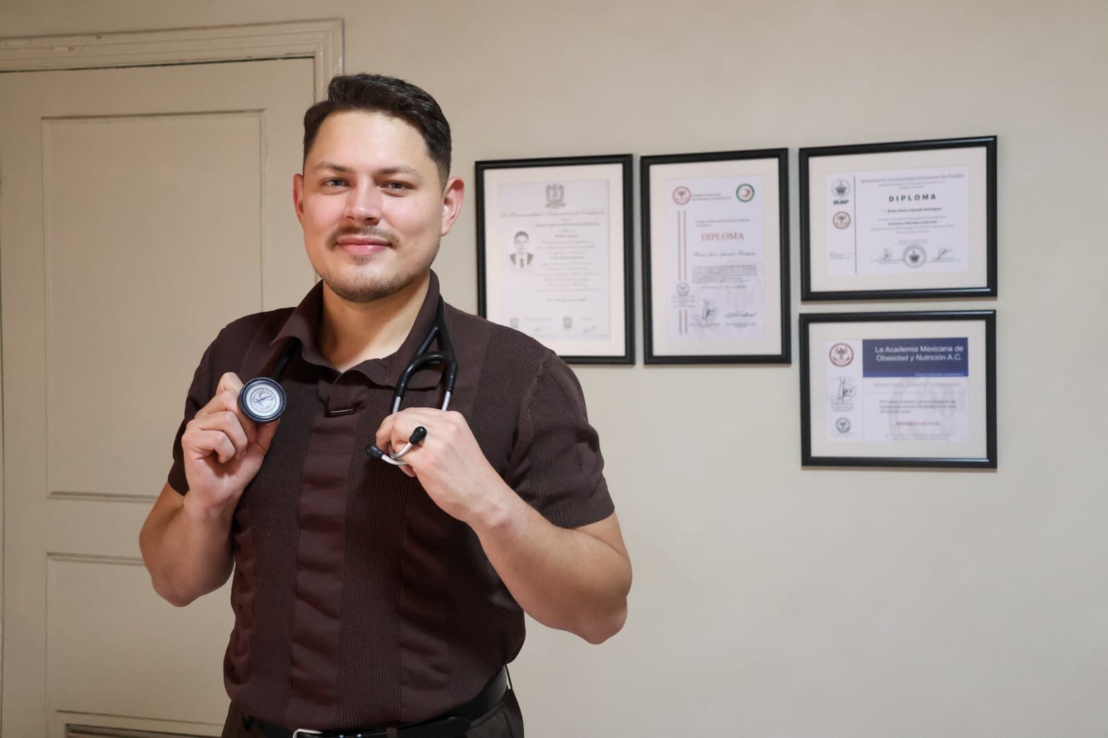 Dr. Brian Amós Zamudio Rodríguez con estetoscopio, frente a los diplomas enmarcados de la BUAP y la Academia Mexicana de Obesidad y Nutrición.